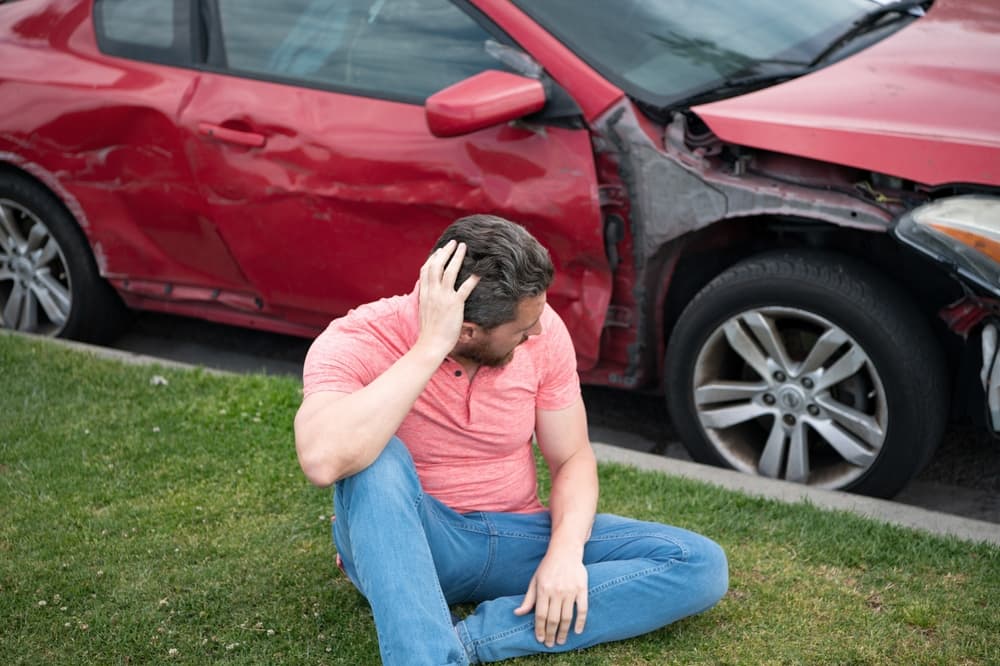 Stunned driver by damaged car after accident. Desperate young man holding head beside broken down car on Chicago road.