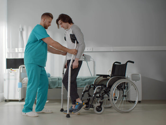 A medical professional assisting a patient with crutches during rehabilitation in a hospital room, with a wheelchair nearby illustrating injuries after a motorcycle accident