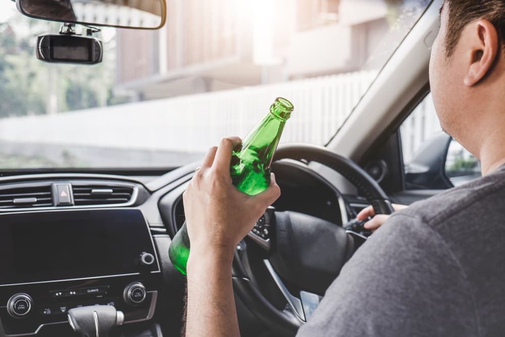 A man driving a car while holding an open bottle of beer behind the wheel.