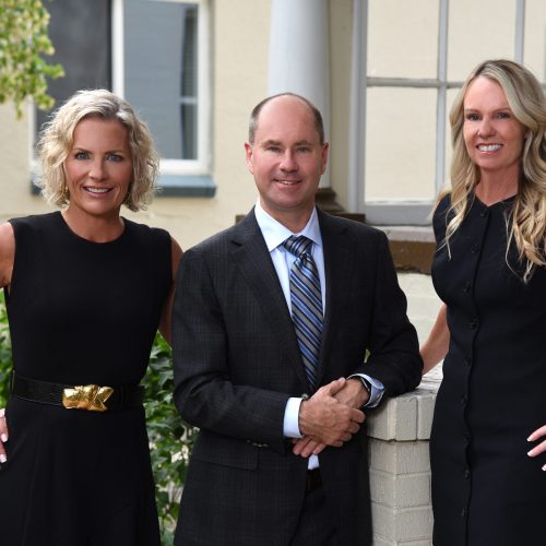 Top Dog Law Sexual Assault Team — two women in black dresses and one man in a dark suit with a striped tie, standing together outside a building, smiling at the camera.